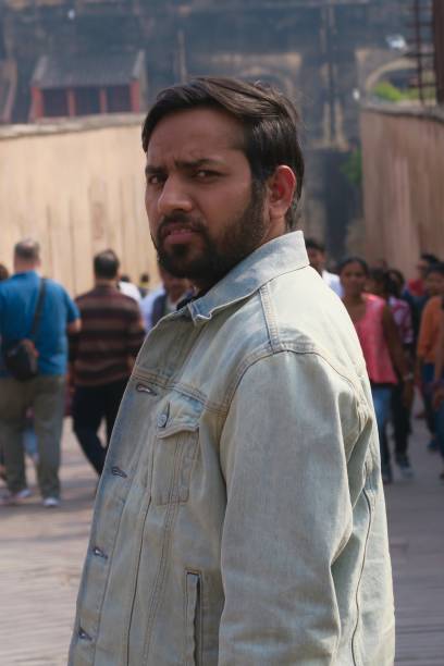 image of man frowning in sunshine, handsome indian man at agra fort with hindu tourists in background photo, tourist crowds on holiday, agra, india - explorer tourist frowning men imagens e fotografias de stock