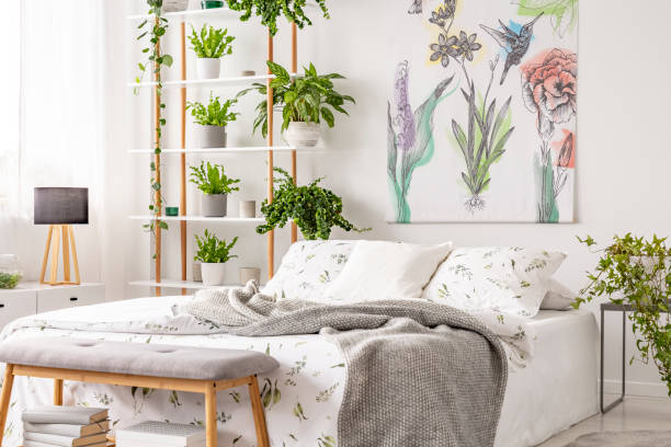 istock Grey blanket thrown on double bed with floral bedclothes standing in white room interior with painting, many fresh plants on wooden rack in the real photo 1146765938