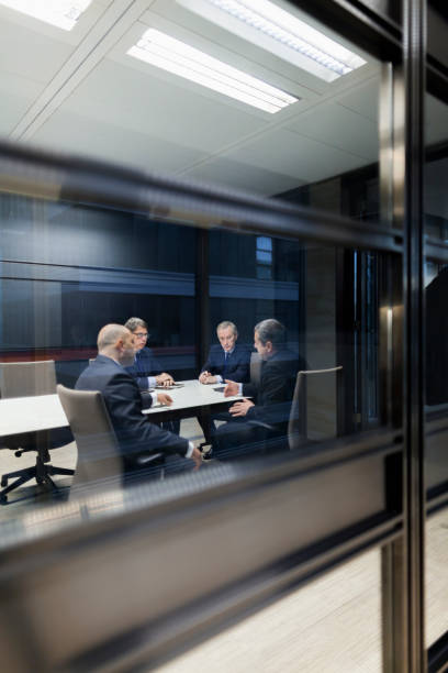Group of businessmen sitting together in meeting View of group of senior adult experienced mature businessmen through glass door reflections in conference meeting room talking strategy ideas and problem solutions privately well dressed suit and tie with leader CEO at head of table old rich white guy stock pictures, royalty-free photos & images