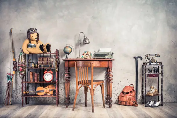 Photo of Vintage old typewriter, lamp, carnival mask, globe on antique table, chair, Teddy Bear with photo camera, retro clock, books, fiddle, keys on shelf, telephone, souvenirs, shoes, cane, backpack, bow