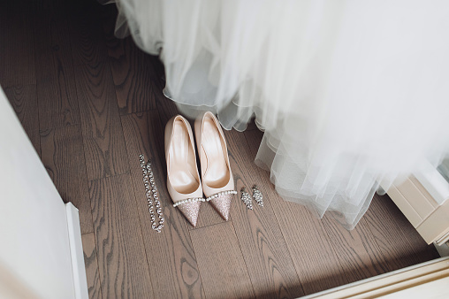 Beautiful pink shoes of the bride stand on the wooden floor near the white dress, golden rings and wedding jewelry. No people.