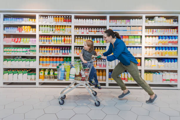 Mother and child racing through supermarket aisle Mother pushing son child shoppers consumers customers in shopping cart in beverages drinks aisle in supermarket grocery store in a hurry racing fast forward chores errands shopping spree the way forward pre press stock pictures, royalty-free photos & images