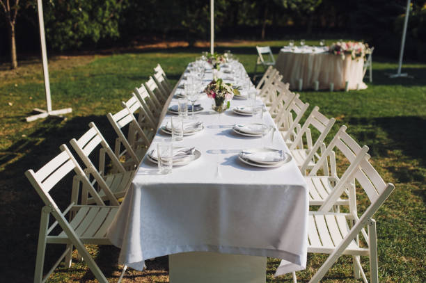 istock Table setting. A large white long table, decorated with fresh flowers, with plates, glasses and forks, stands in a park with green grass. Preparing for a wedding party outdoors.. 1146782881
