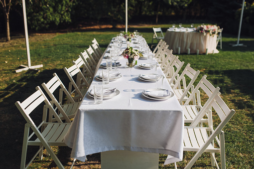 A large white long table, decorated with fresh flowers, with plates, glasses and forks, stands in a park with green grass. Wedding decorations and details. Preparing for a wedding party.
