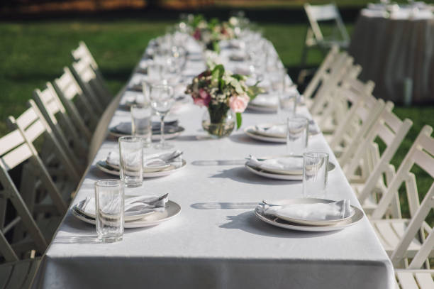 istock Table setting. A large white long table, decorated with flowers, with plates, glasses and forks, stands in a park with green grass. Wedding decorations and details. Wedding party outdoors. 1146782883