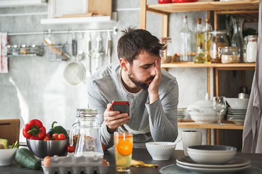 Front view of a young man looking sad while using smart phone at home. He is having breakfast at the kitchen counter.