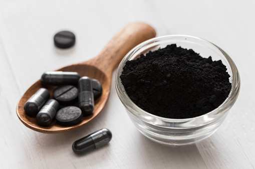 Activated charcoal powder in a glass bowl and pills on a spoon on white wooden background