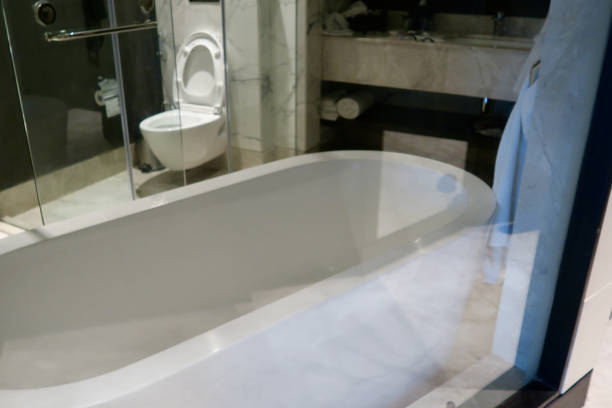 Stock photo showing a white luxury bathroom suite, laid out with good home interior design. The washroom features oval bath, marble tiles, glass wall to bedroom, wall mounted toilet. imagen de baño de lujo blanco suite diseño interior con baño ovalado, azulejos de mármol caros, dormitorio en suite baño de pared de vidrio piso a techo ventana, baño independiente, grifo montado en el suelo cascada, cabina de ducha, wc montado en la
