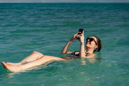 woman taking a tan and relaxing in the dead sea. Checking social media with the phone