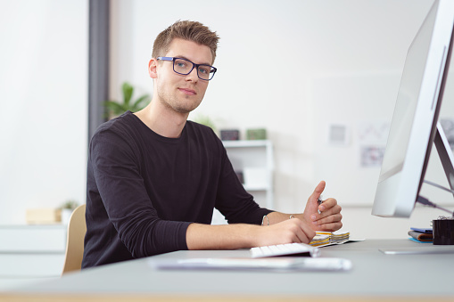 Single handsome young man wearing eyeglasses at computer on desk in bright office with white walls