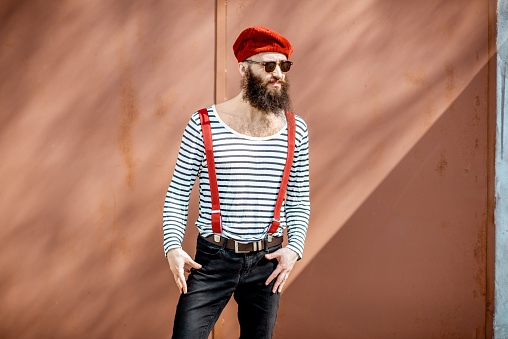 Portrait of a stylish bearded man dressed in striped shirt, suspenders and hat on the red background outdoors