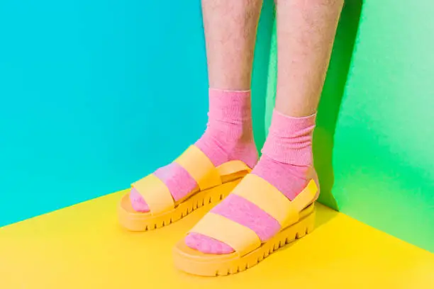 Male hairy legs in socks staying in women's sandals on bold background in the corner with strong shadows. Minimal pride concept. Body part