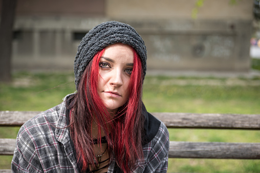 Homeless girl, Young beautiful red hair girl sitting alone outdoors on the wooden bench on the street with hat and shirt feeling anxious and depressed after she became a homeless person