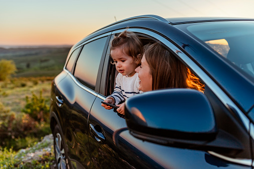 Family vacation theme. Young woman with kid looking out from car window during a road trip in summer. Photo of happy Caucasian mother and son sticking heads out of the window of a car looking at the scenery.