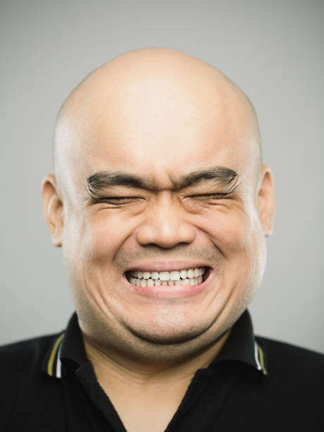 Portrait of real chinese adult man with extreme expression and eyes closed Close up portrait of asian adult man with shocked expression and eyes closed against gray white background. Vertical shot of chinese real people with extreme positive or negative emotion in studio with bald head. Photography from a DSLR camera. Sharp focus on eyes. clenching teeth stock pictures, royalty-free photos & images