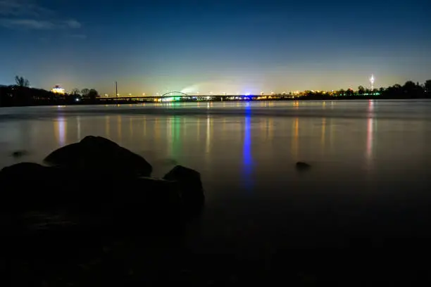 Reflections of the city lights of Düsseldorf and Neuss in the river Rhine at night. The cities are connected by a bridge. Rocks in the foreground - long exposure, blue mood