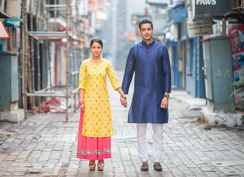 An Indian couple stands in the middle of the street outside. They are beautifully dressed.