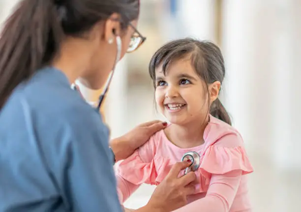 A toddler girl sits on a table during a medical examination. A female medical professional is using a stethoscope to listen to her heartbeat. The child is smiling up at her doctor.