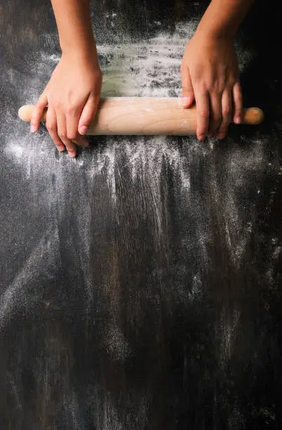 Photo of Girl hands keep rolling pin with flour on dark black table, baking background, top view, copy space for your text, meru, recipe. Banner