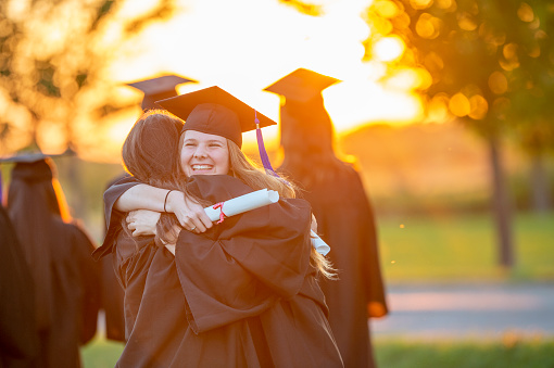 A blonde woman hugs a graduating student wearing a mortarboard, while the sun sets in the background.