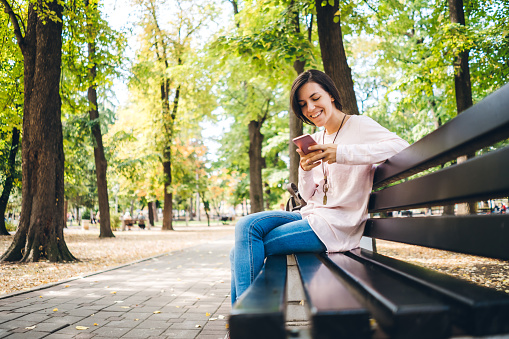 Young woman sitting on the beach in public park, using mobile phone, smiling, enjoying the day.