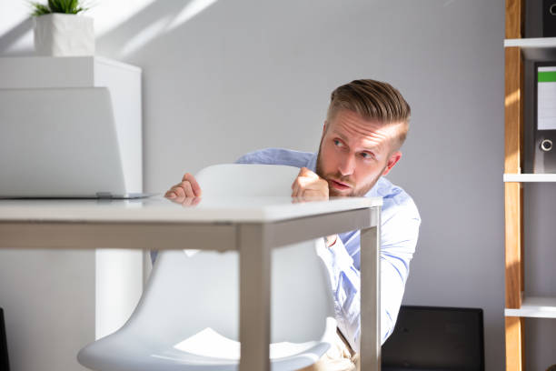 jeune homme d’affaires effrayé se cachant derrière la chaise au lieu de travail - hiding fear business men photos et images de collection