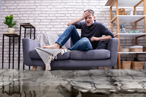 Upset Young Man Sitting On Sofa In The Living Room Flooded With Water