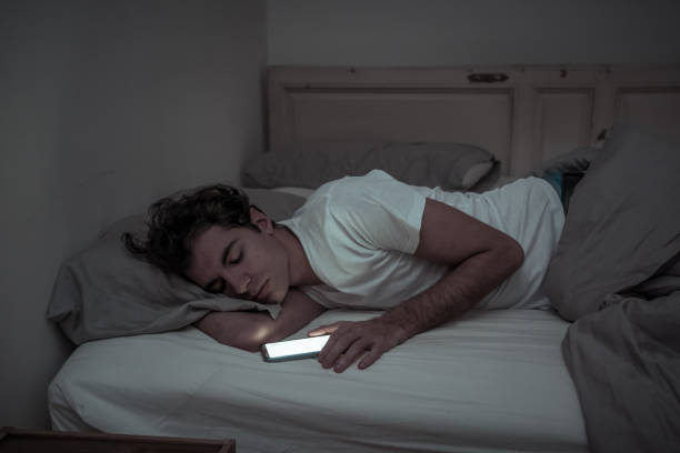 Addicted to social media young man falling asleep with smart mobile phone at night in bed. lifestyle portrait of man sleeping in dark bedroom with mobile screen light on. Mobile use addiction. stock photo