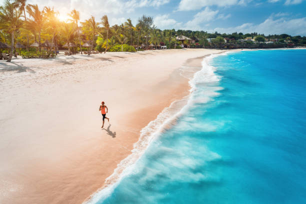 luftaufnahme der laufenden jungen frau am weißen sandstrand nahe dem meer mit wellen bei sonnenuntergang. sommerferien. top ansicht des sportlich schlanken mädchens, klares azurblaues wasser. indischer ozean. lifestyle und sport - zanzibar stock-fotos und bilder