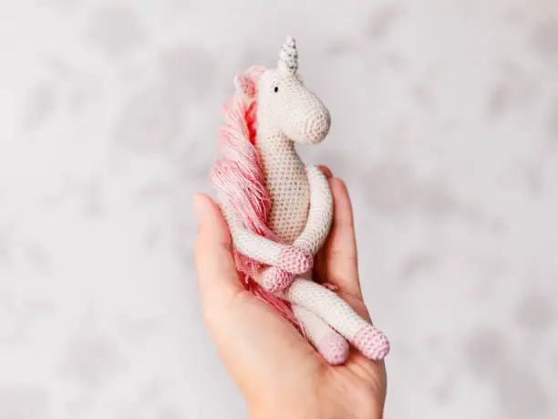 Cute fairy unicorn with a pink mane and a tail made of threads. Crocheted hand made toy on woman palm. Trendy creature, symbol of magic and miracles.