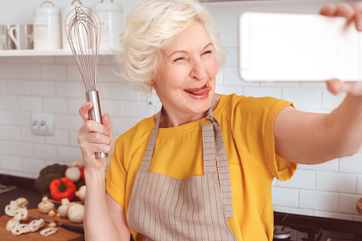 Handsome grandmother makes funny selfie with a whisk in the kitchen.