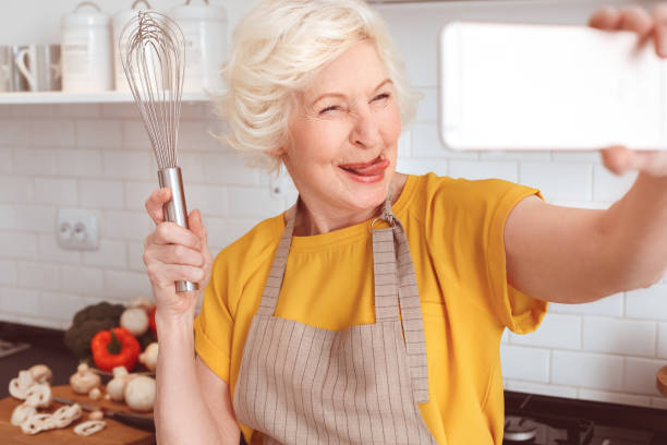 Handsome grandmother makes funny selfie with a whisk in the kitchen. Tongue out, crazy face. Handsome grandmother makes funny selfie with a whisk in the kitchen. Indoor, studio shoot, kitchen interior sticking out tongue photos stock pictures, royalty-free photos & images