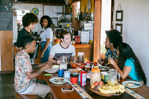 Candid view of Brazilian friends sitting around table with people in kitchen preparing food and drink, backpackers, gap year students