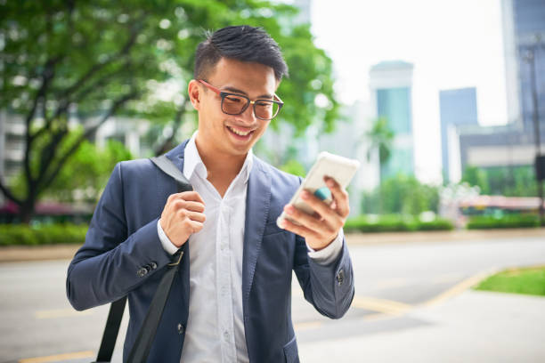 Young malaysian businessman on smart phone Young malaysian businessman in the streets of downtown Kuala Lumpur smiling while looking to his his smart phone for a crowed sourced taxi to arrive . Kuala Lumpur, Malaysia, Asia malaysia office workers stock pictures, royalty-free photos & images