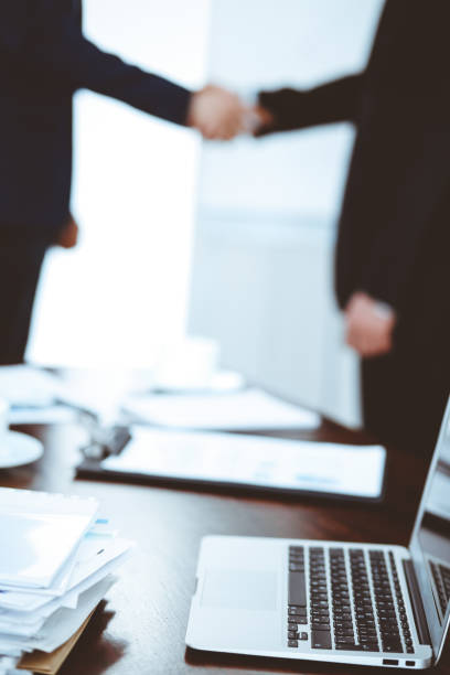 istock Documents and laptop on the table. Business people shaking hands on the background, silhouettes 1147053048