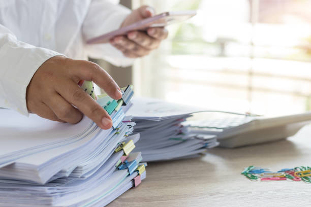 istock Teacher hand is holding smartphone and searching student report and homework assignment on desk for score. Unfinished paperwork stacked in archive with color paper clip. Education and business concept 1147069725