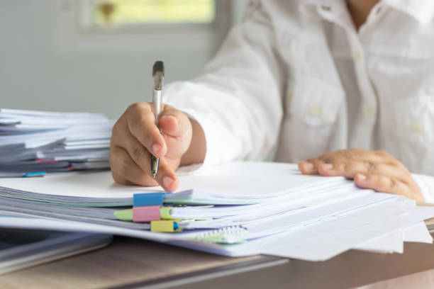 la mano del maestro está sosteniendo la pluma para revisar las tareas escolares del estudiante en el escritorio en la escuela. papeleo inconcluso apilado en archivo con papel color y clips de papel aglutinante. el concepto educativo y empresarial - paper clip clip paper office fotografías e imágenes de stock