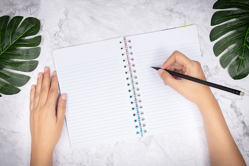 flat lay of woman hand writing in a blank white page notebook on white marble desk, top view. green palm leaf on desk for decoration