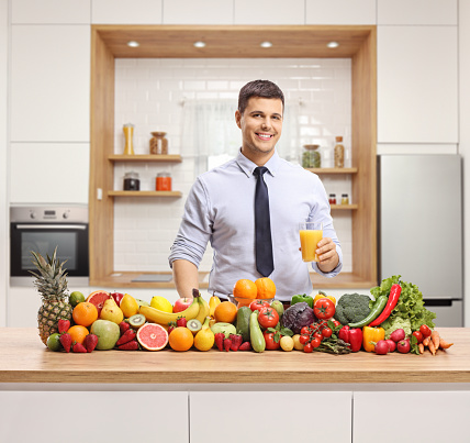 Young man posing with a glass of orange juice and a pile of fruits and vegetables on a wooden counter in a kitchen