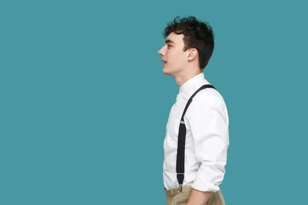 Profile side view portrait of happy handsome hipster curly young businessman in classic casual white shirt and suspender standing and looking straight. indoor studio shot isolated on blue background.