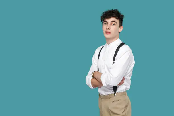 Profile side view portrait of satisfied handsome hipster curly young businessman in classic white shirt, suspender standing, crossed arms and looking at camera. studio shot isolated on blue background