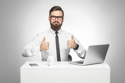 Everything alright! Portrait of handsome satisfied bearded young businessman in white shirt and black tie are sitting in office and working on laptop with smile and showing thumb up, looking at camera