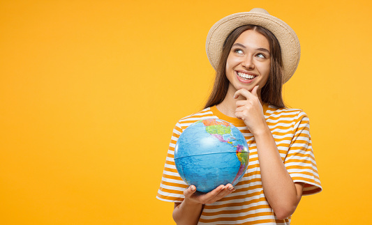 Young female dreaming about future trip, holding globe with one hand, isolated on yellow background. Travel concept.
