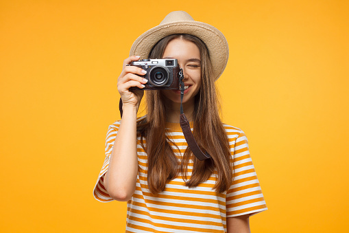 Smiling young girl tourist holding camera, isolated on yellow background