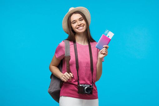 Excited young woman tourist with beautiful smile holding passport with tickets, isolated on blue background
