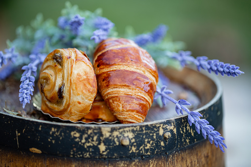 French croissant and bun with raisins on old wine barrel. Dessert. Bouquet of lavender flowers. Dinner in a garden in summertime. Summer twilight. Soft focus. Blurry background. Close-up. Macro.