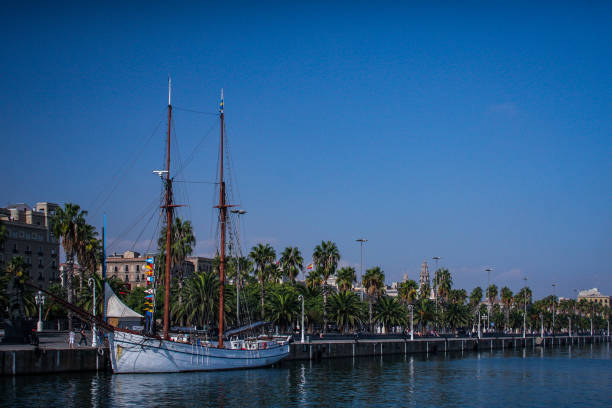 An old style looking ketch white sailboat is moored in the beachfront of Barcelona, Spain, on a clear sunny day without clouds. Palms and houses are seen behind the sailboat.