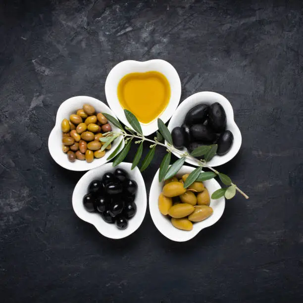 still life with ceramic bowls, in the shape of a heart, with extra virgin olive oil and different types of olives, on a rough black background. Top view