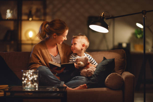 familie voor het slapen gaan moeder leest haar kind zoon boek in de buurt van een lamp in de avond - lezen fotos stockfoto's en -beelden