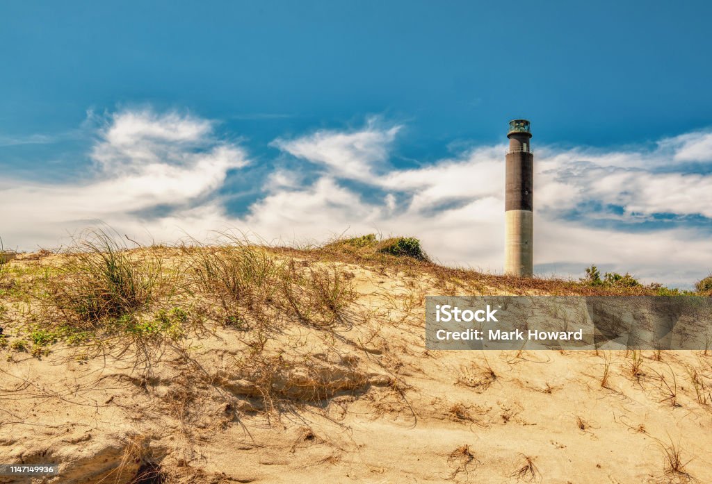 Lighthouse On The Hill A slender black and white beach lighthouse above a sandy dune with a cloudy blue sky. Oak Island - North Carolina Stock Photo